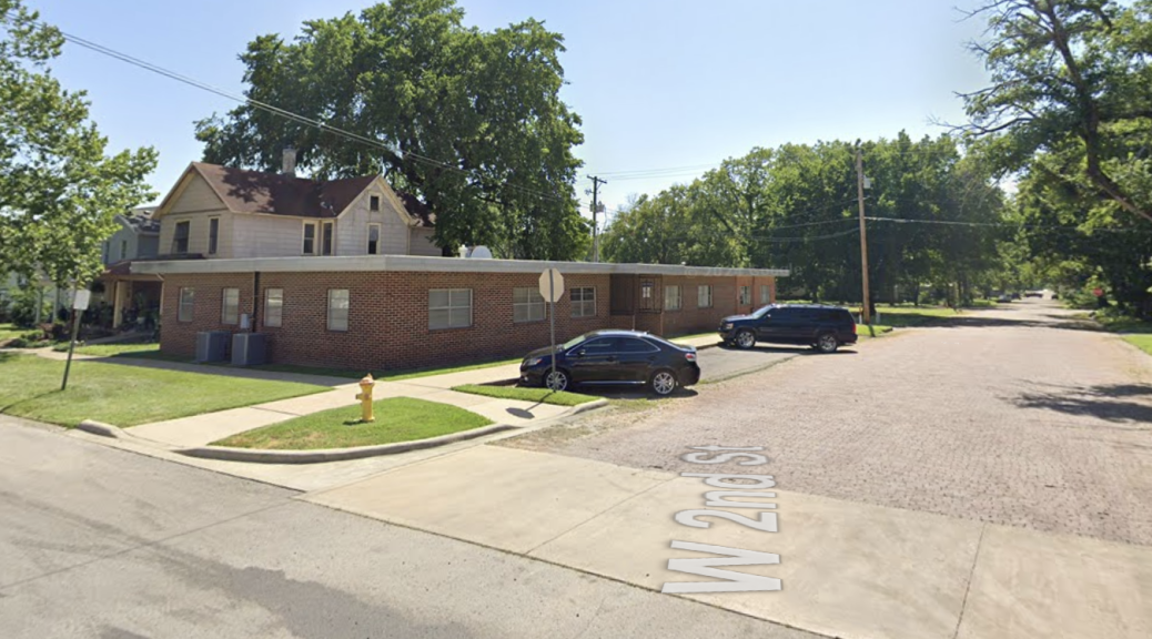 A single-story brick building on the corner of West Second Street in Fort Scott, with a two-story house behind it and cars parked at the curb.