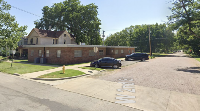 A single-story brick building on the corner of West Second Street in Fort Scott, with a two-story house behind it and cars parked at the curb.