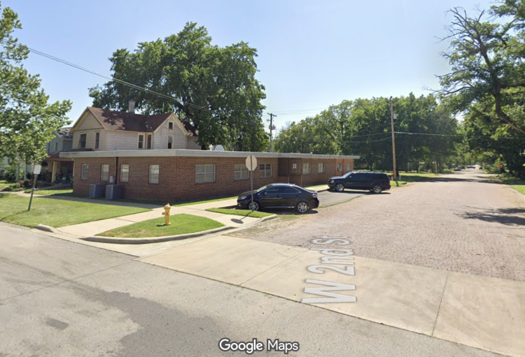 A single-story brick building on the corner of West Second Street in Fort Scott, with a two-story house behind it and cars parked at the curb.